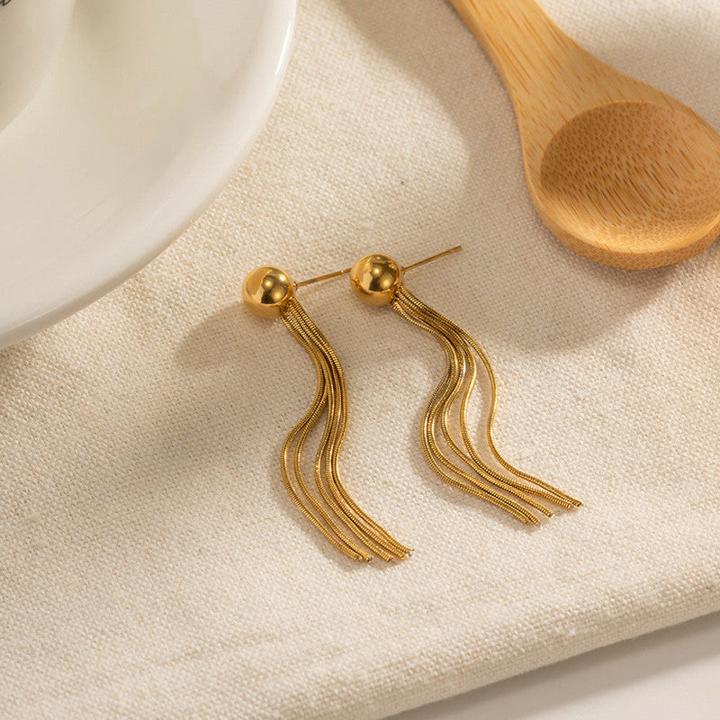 Gold tassel earrings on a beige fabric background with a wooden spoon and ceramic dish.