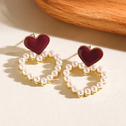Pair of earrings with red heart-shaped tops and pearl-studded gold loops on a light background.